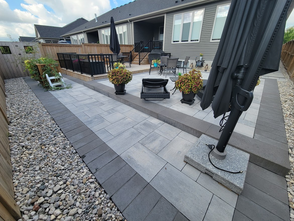 A modern backyard patio with gray tiles, potted plants, outdoor furniture, a large umbrella, and a surrounding wooden fence on a sunny day.