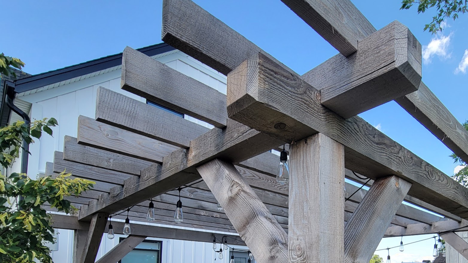 A rustic wooden pergola with hanging lights stands adjacent to a white building under a clear blue sky with some greenery visible.