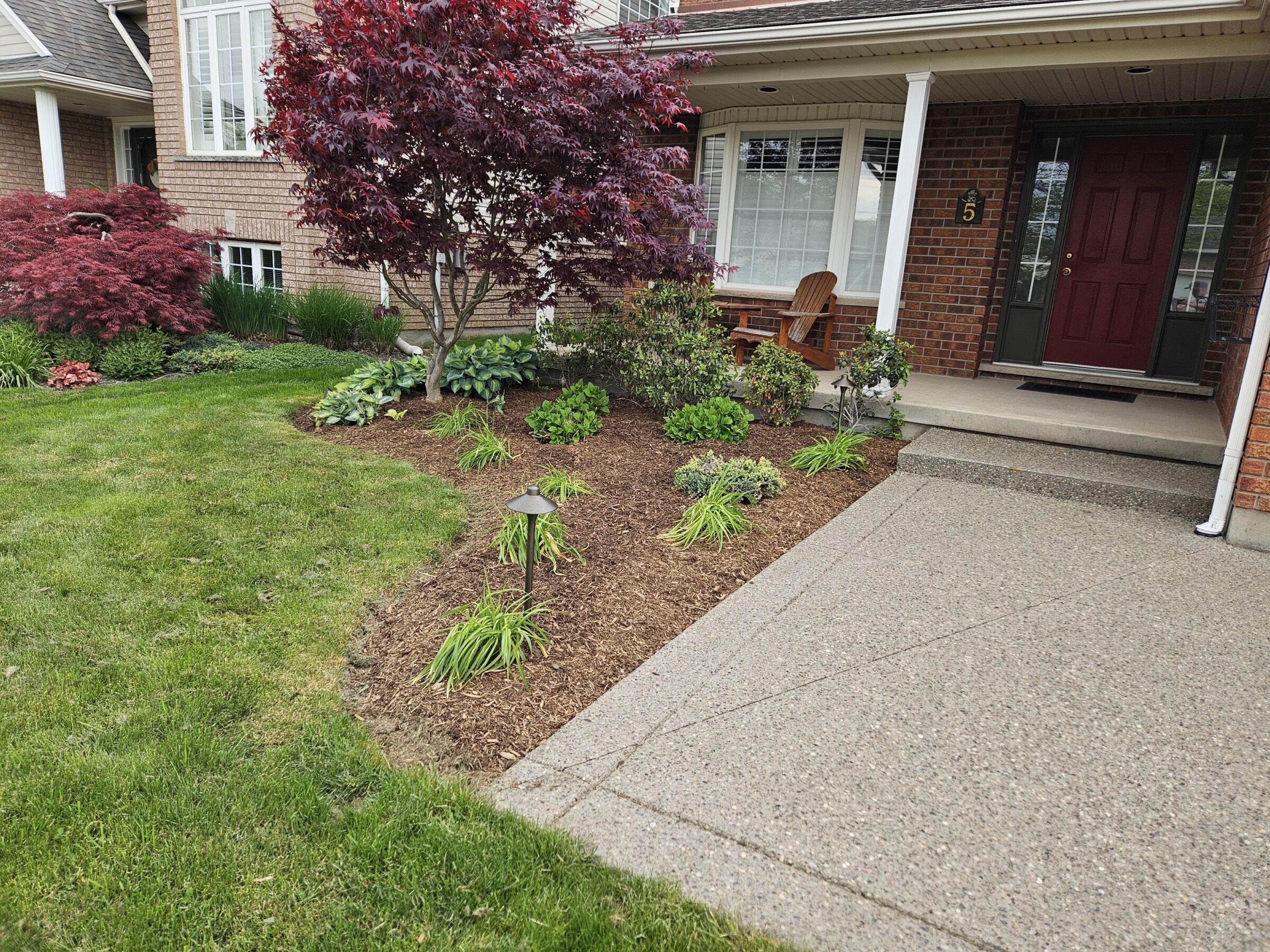 A well-maintained front yard with a red door, landscaped garden beds, a concrete walkway, and a wooden chair on the porch.