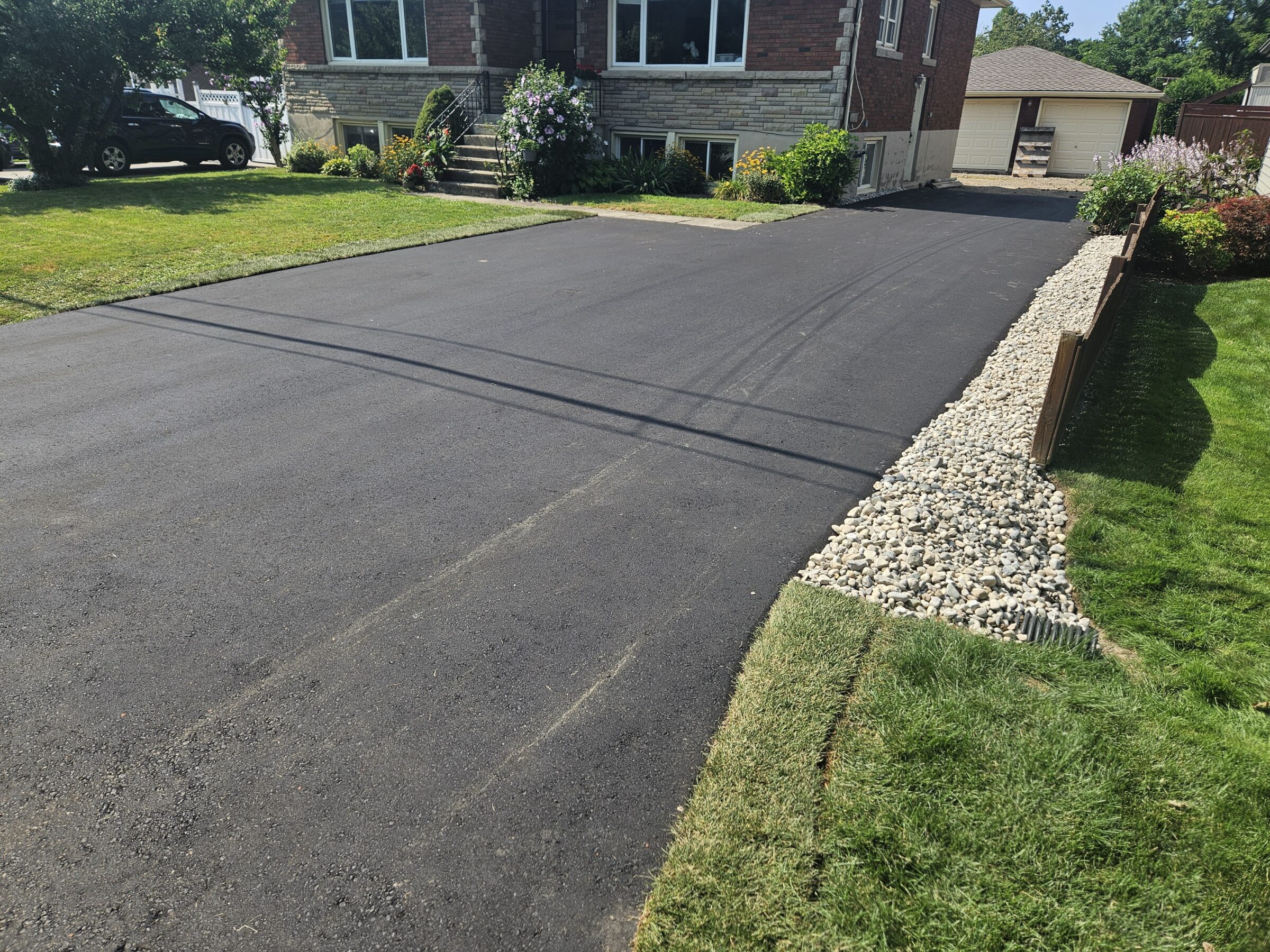 A freshly paved driveway leads to a brick house with a lush garden and garage in a quiet, suburban neighborhood.