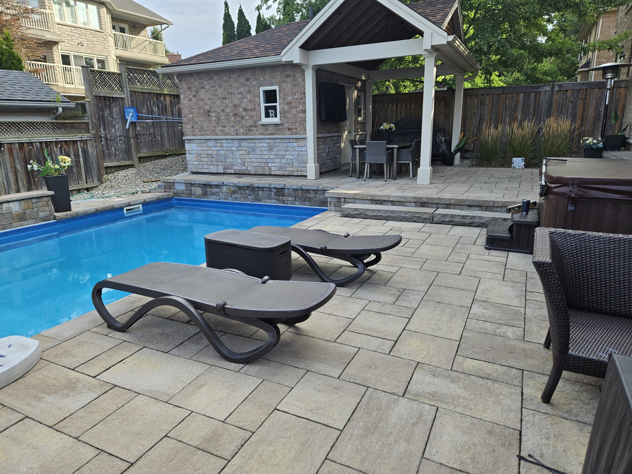 A backyard pool area with lounge chairs, a gazebo, outdoor dining set, and privacy fence. Nearby houses are visible in the background.