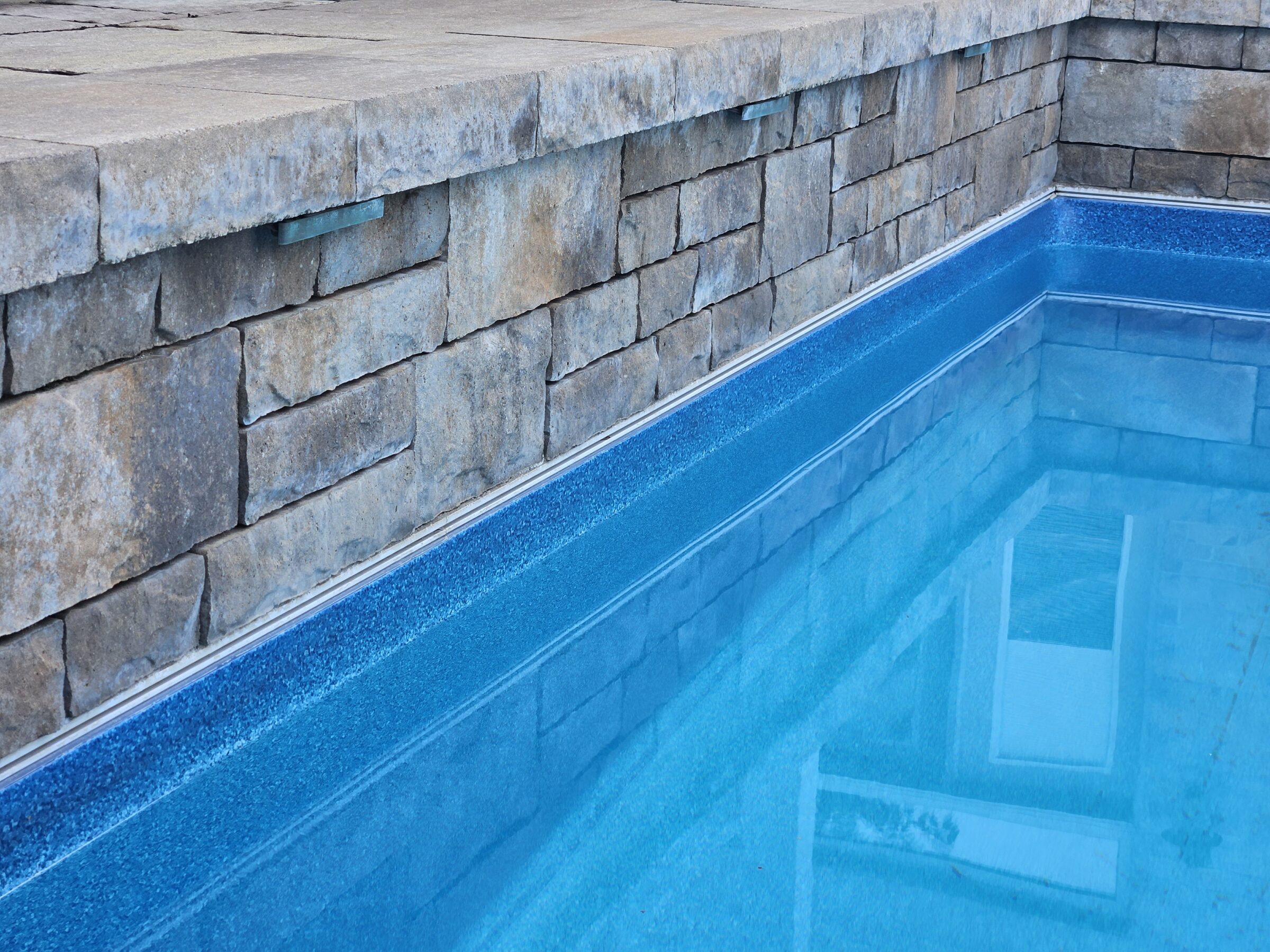 A close-up of a stone wall by an empty swimming pool, with clear blue water reflecting part of a building.