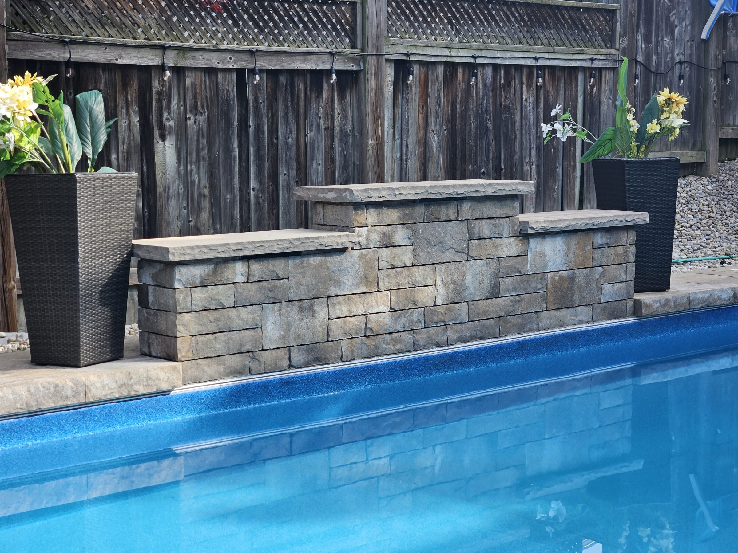 A backyard pool with a stone bench, plastic wicker planters with flowers, and a wooden fence adorned with string lights in the background.