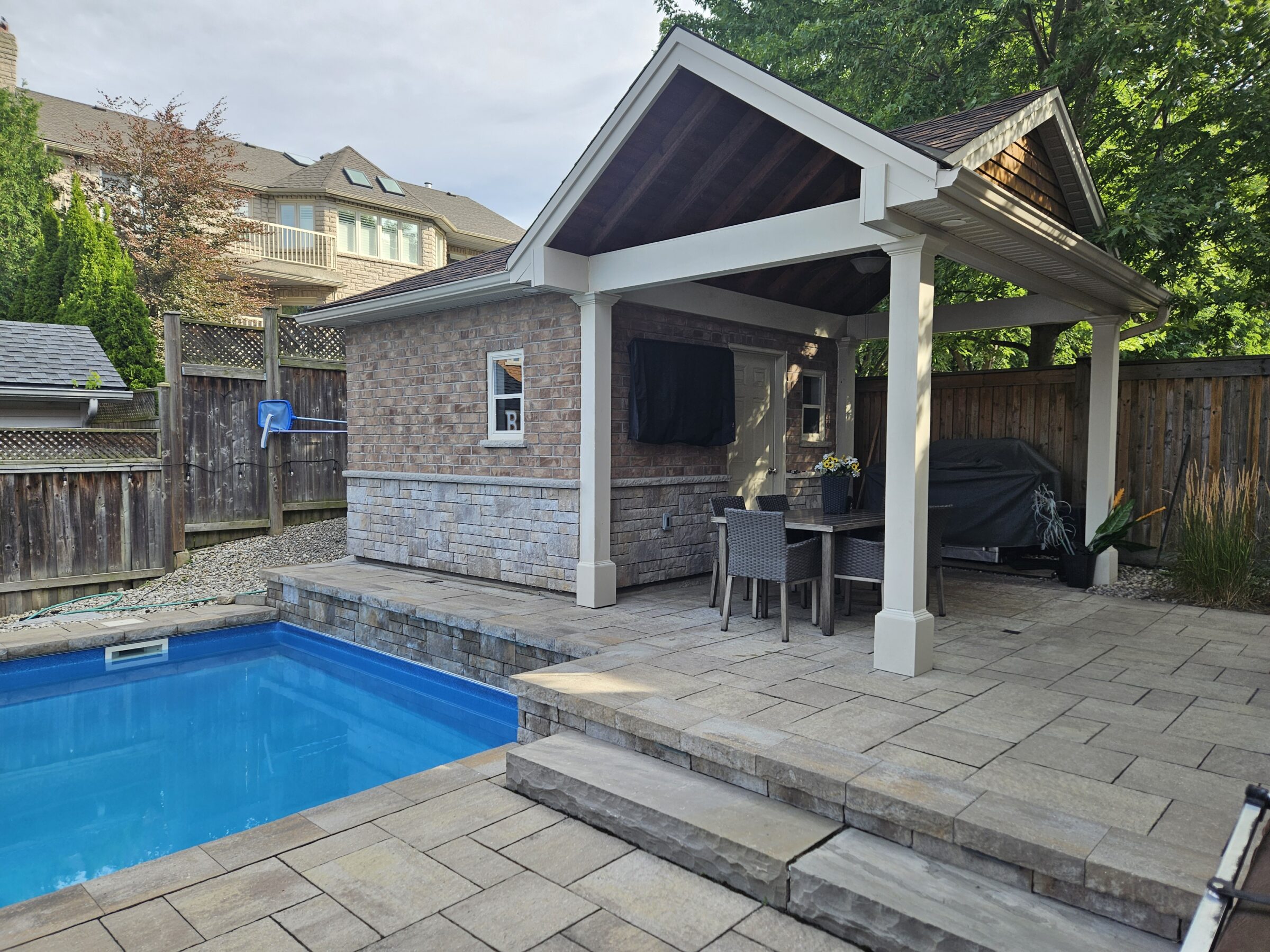 Backyard with a pool, fenced area, patio, and covered seating. Trees and neighboring house visible. Relaxed residential outdoor setting.