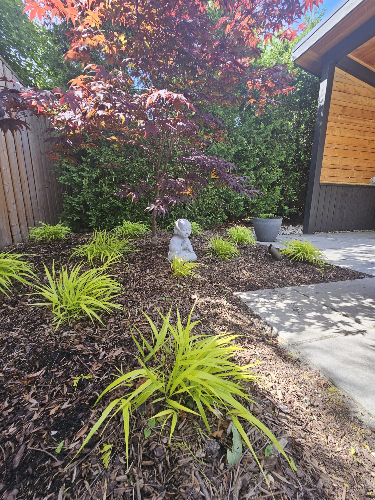 A serene garden with a small statue, vibrant foliage, and a wooden structure on the side. No recognizable landmarks or historical buildings.