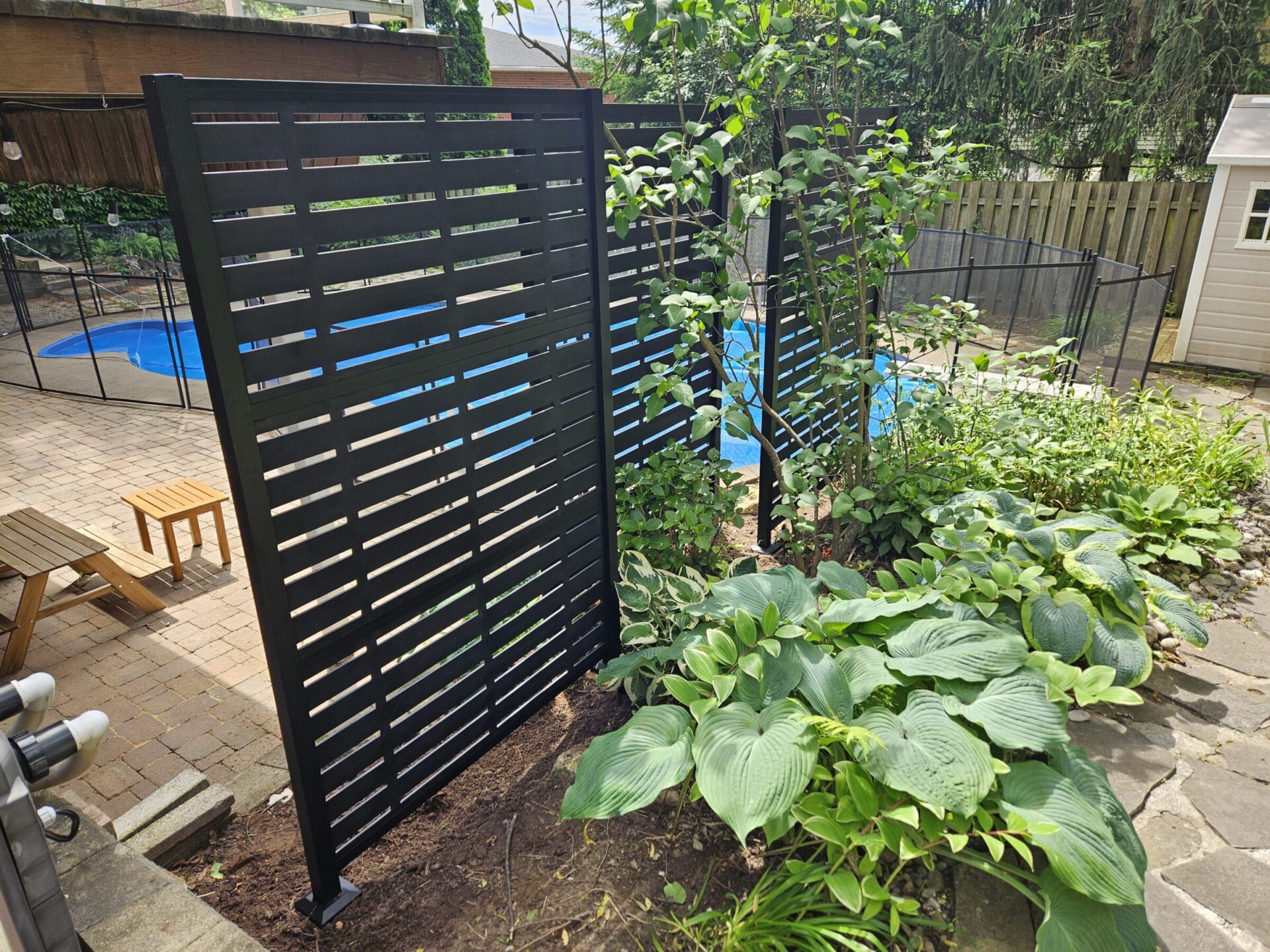 A backyard features a black slatted screen, green plants, a swimming pool with safety fence, and a picnic table on a brick patio.