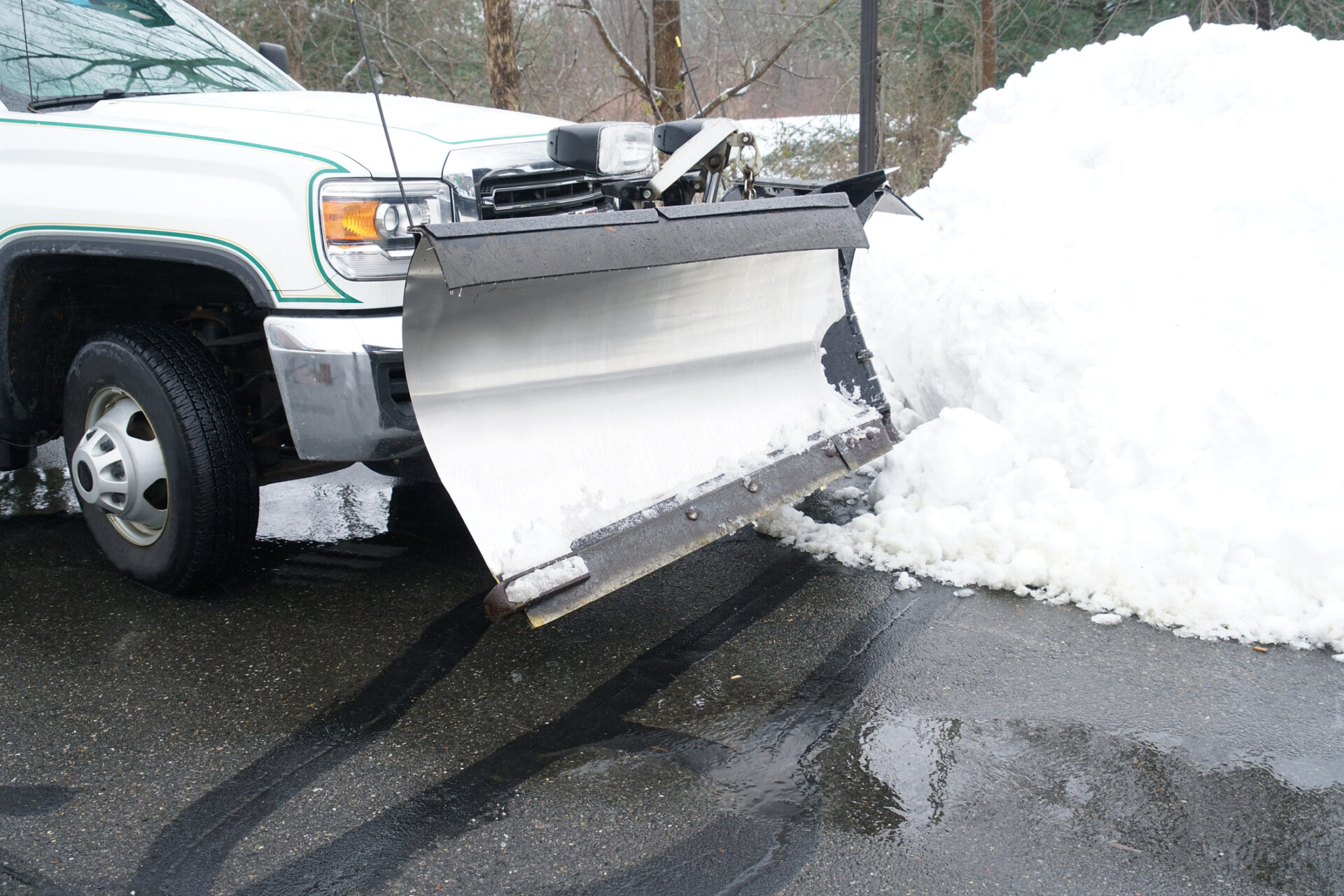 A truck with a snowplow attachment clears a snow-covered road, leaving wet, slushy tracks behind. Trees and snow are visible in the background.