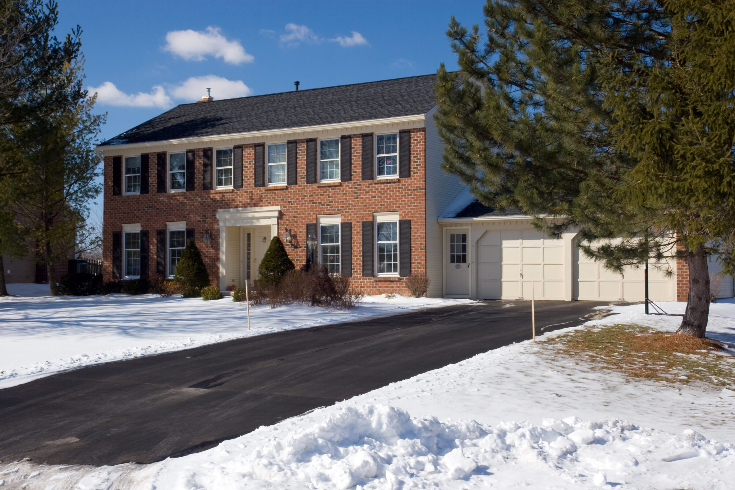 A two-story red brick house with snow-covered yard, tall trees, black shutters, and a double garage under a blue sky.