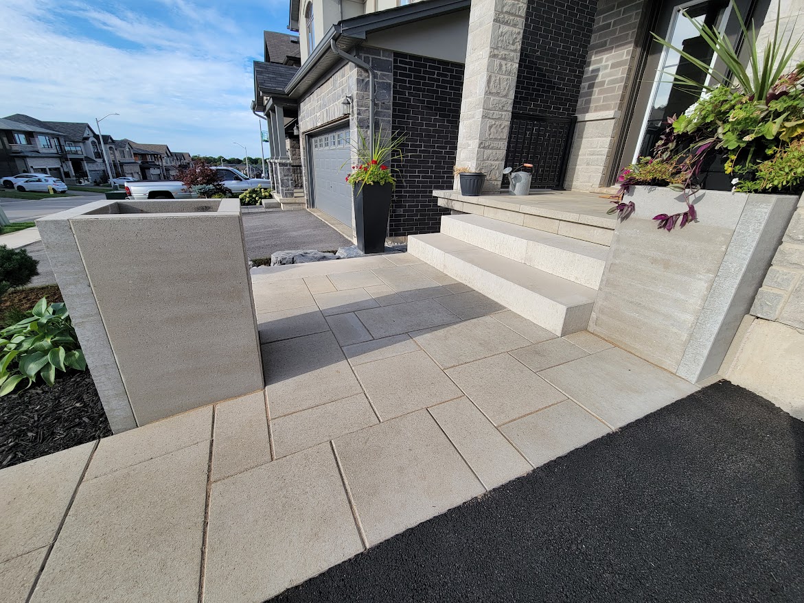 Modern suburban house with stone steps, planters, and a driveway. Neatly arranged plants and houses in the background under a bright blue sky.