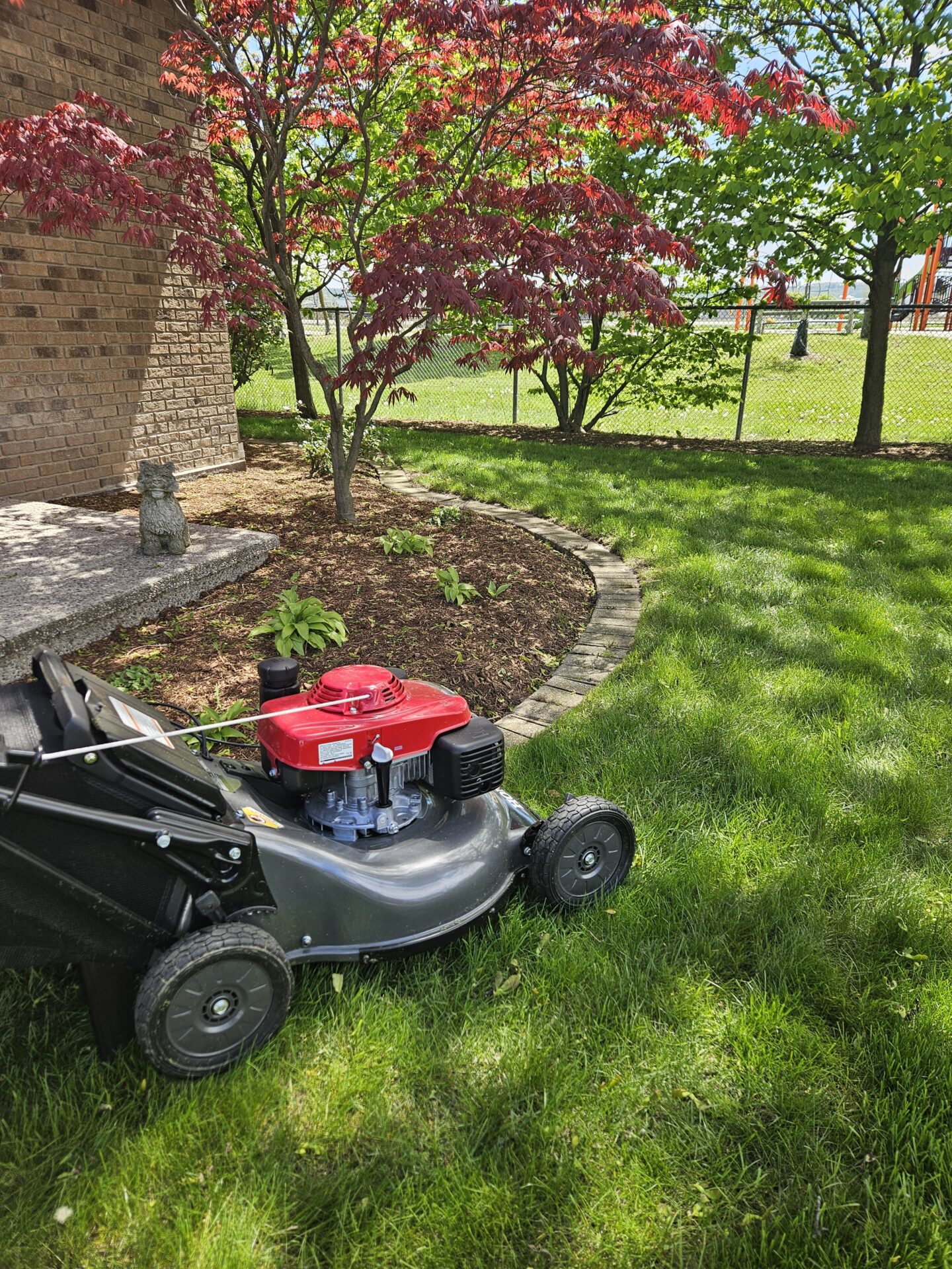 A lawnmower on green grass near a tree with red leaves, beside a brick building and a chain-link fence with a playground in the background.