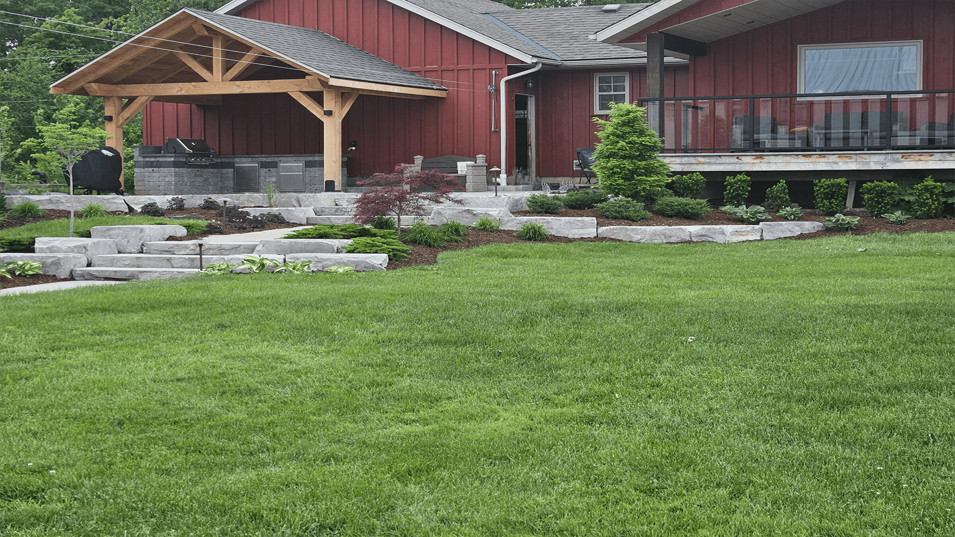 A well-maintained garden with neatly cut grass, stone steps, various plants, and a red wooden house featuring an outdoor covered kitchen area.