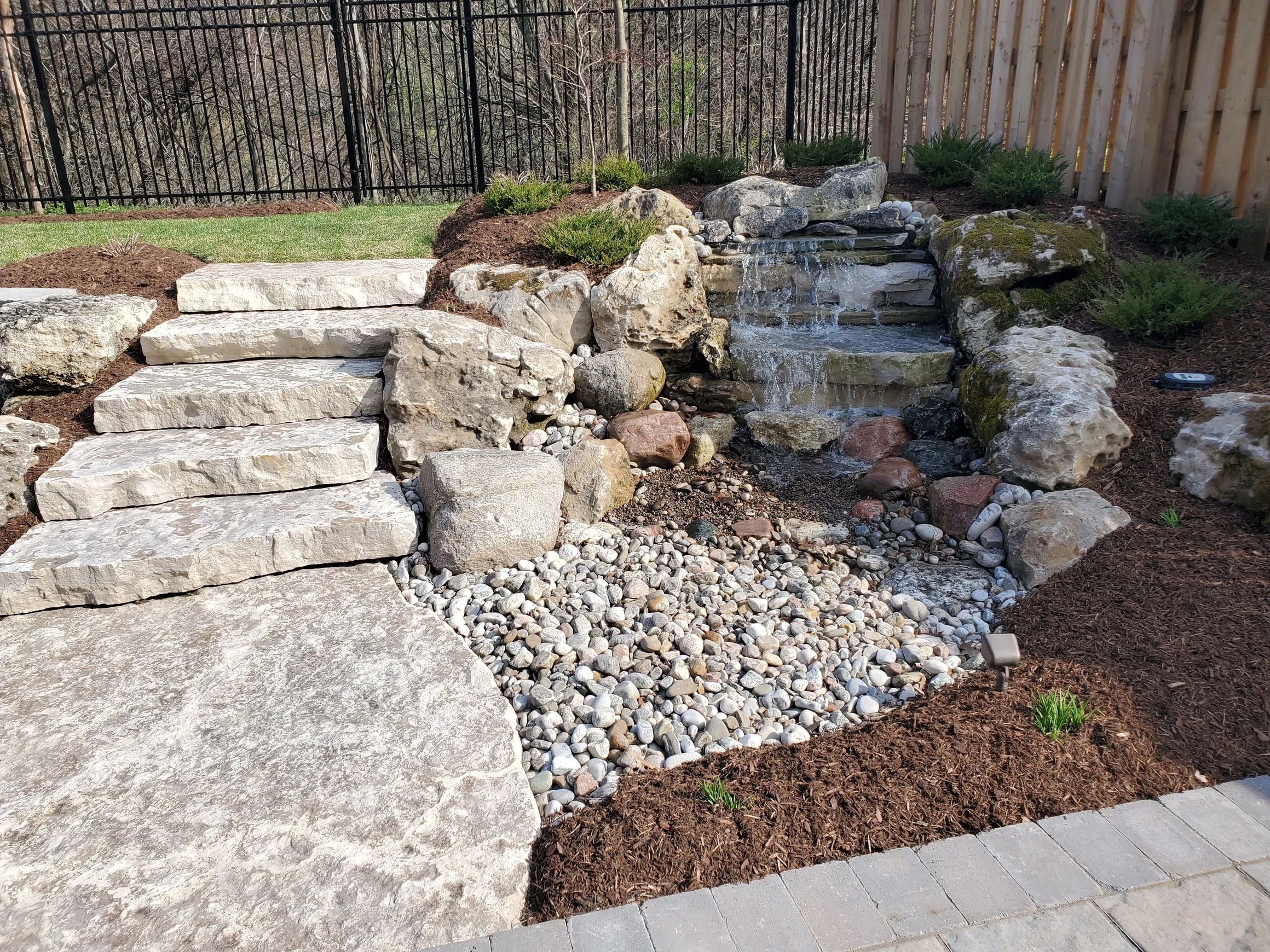 A serene garden features stone steps, a small waterfall, and a pebble stream, bordered by mulch and greenery, with a black metal fence behind.