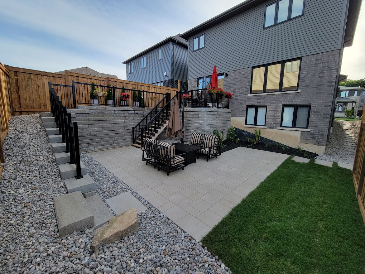 Modern backyard with a spacious patio, outdoor furniture, and stone landscaping, adjacent to a two-story house featuring gray brick and wooden fencing.