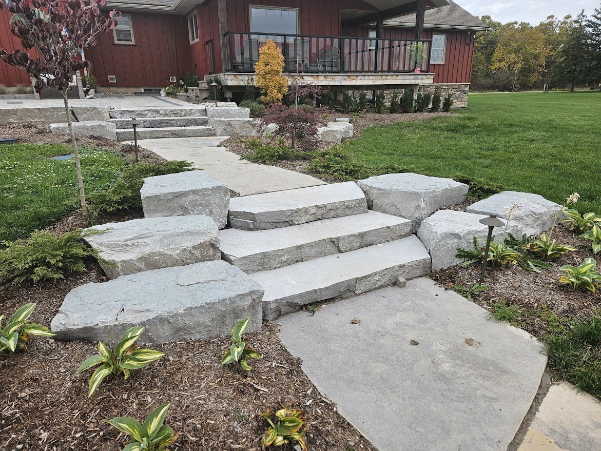 A red house with a stone walkway, steps, and landscaped garden featuring small plants and shrubs, leading to a porch with chairs.