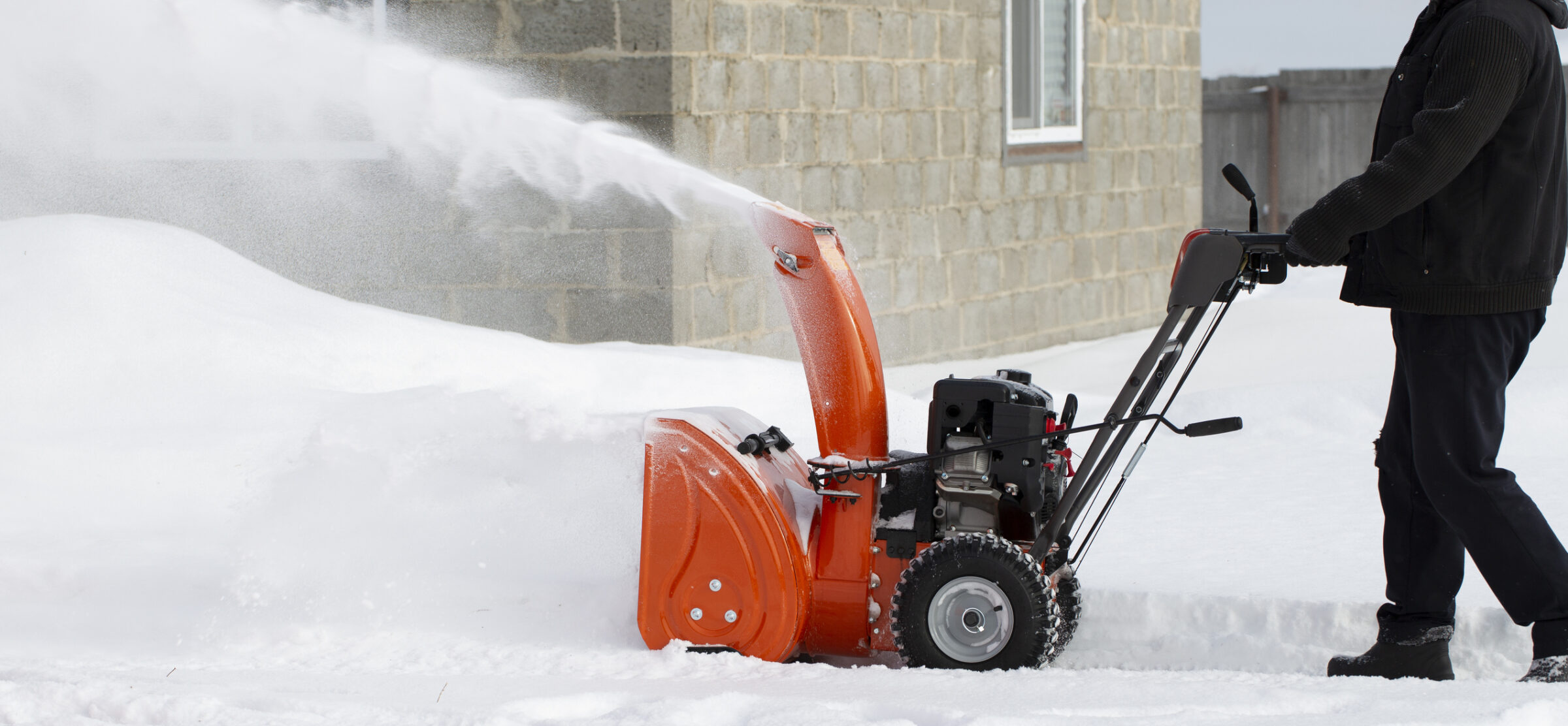 A person operates an orange snowblower, clearing a path of deep snow in front of a brick building on a winter day.