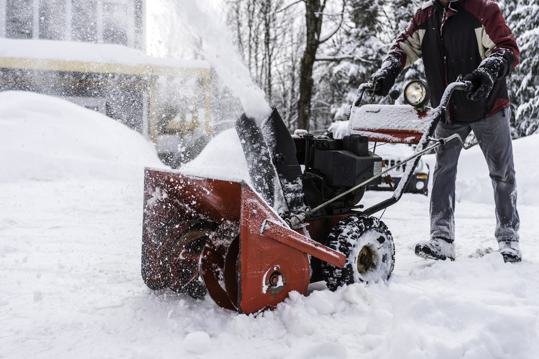 A person operates a snowblower, clearing snow in a snowy landscape, with trees and a building in the background.