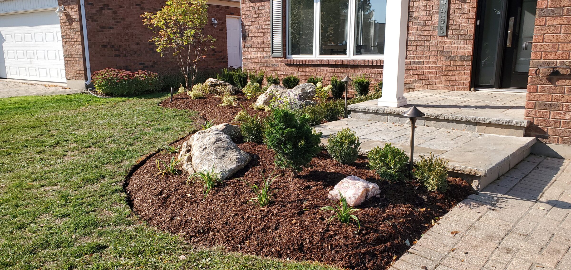 A front yard with landscaped garden, featuring rocks, small shrubs, mulch, and a brick house with a stone pathway and porch.