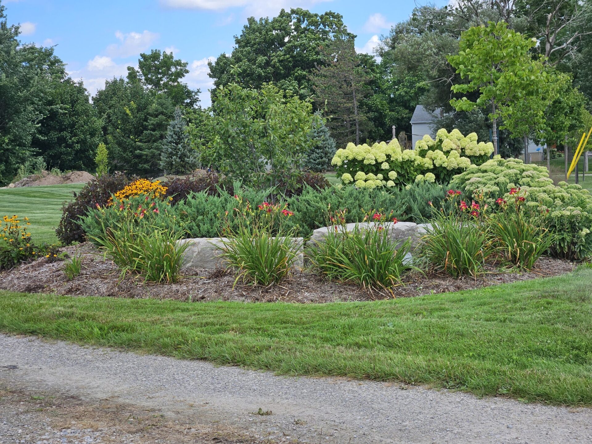 A colorful flower bed with various plants in a well-maintained park setting, bordered by trees and a gravel path on a sunny day.