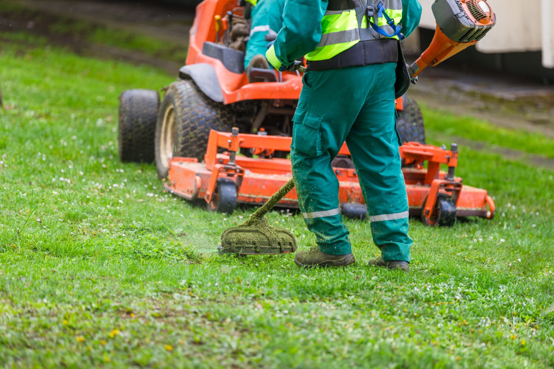 A person in green workwear operates a string trimmer, with an orange lawnmower in the background, cutting grass on a green lawn.