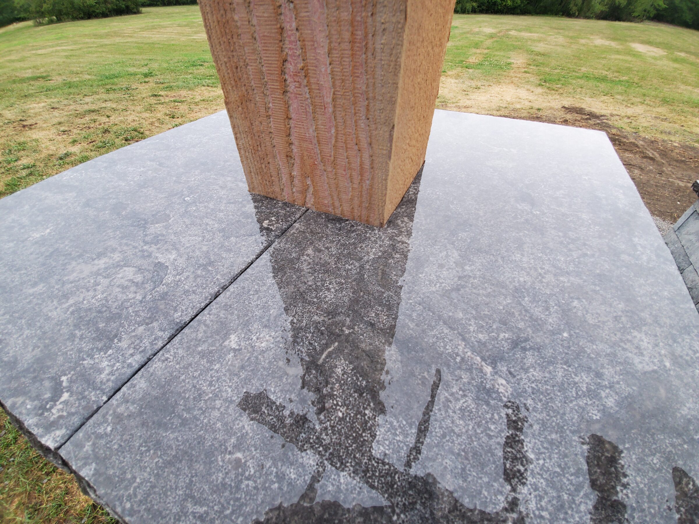 A person’s reflection is visible on a stone surface around a vertical structure in a grassy area with surrounding trees.