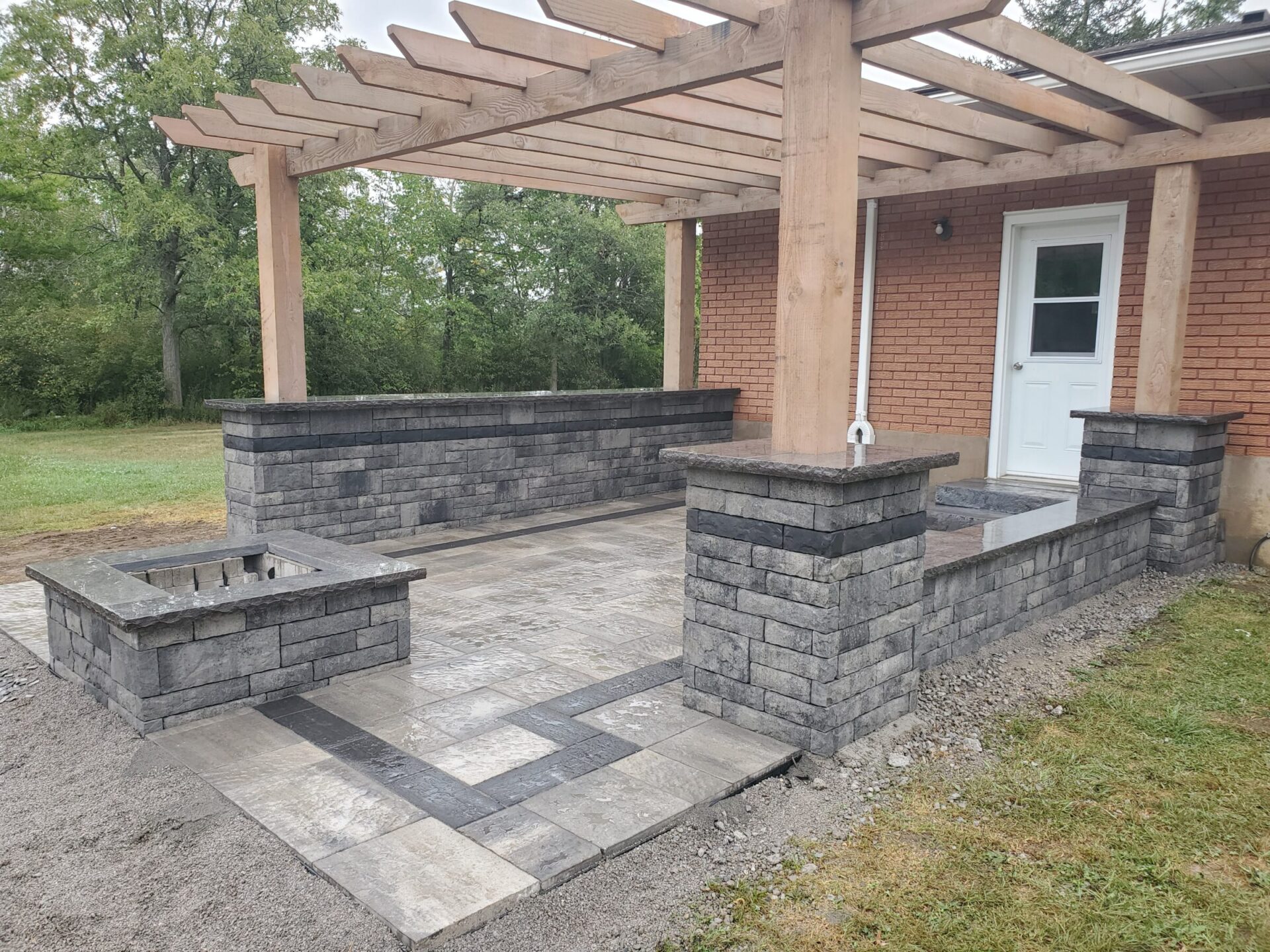 A brick house with a wooden pergola and stone patio, set against a backdrop of green trees and grassy area.